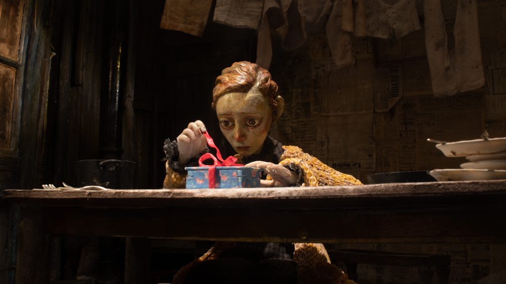 A puppet with short hair and a worried expression sits at a wooden table, untying a red ribbon on a blue gift box