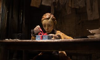 A puppet with short hair and a worried expression sits at a wooden table, untying a red ribbon on a blue gift box