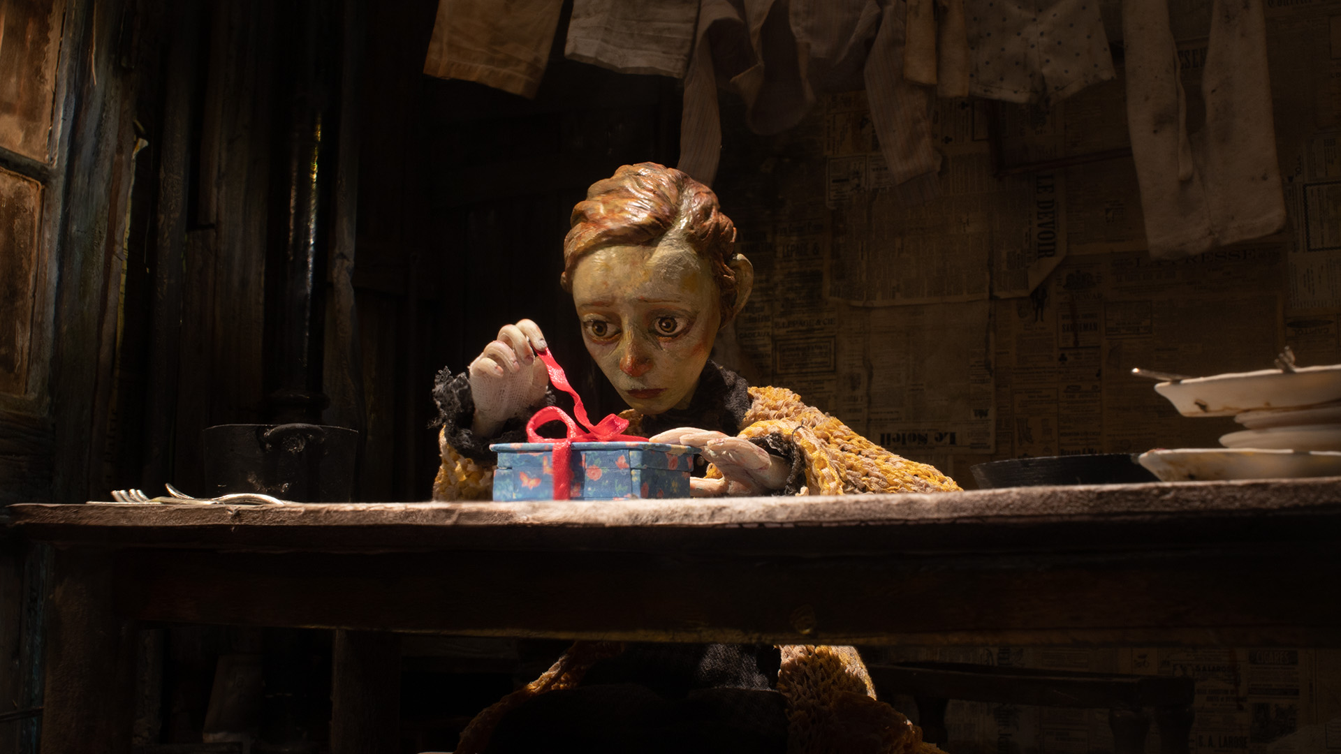 A puppet with short hair and a worried expression sits at a wooden table, untying a red ribbon on a blue gift box