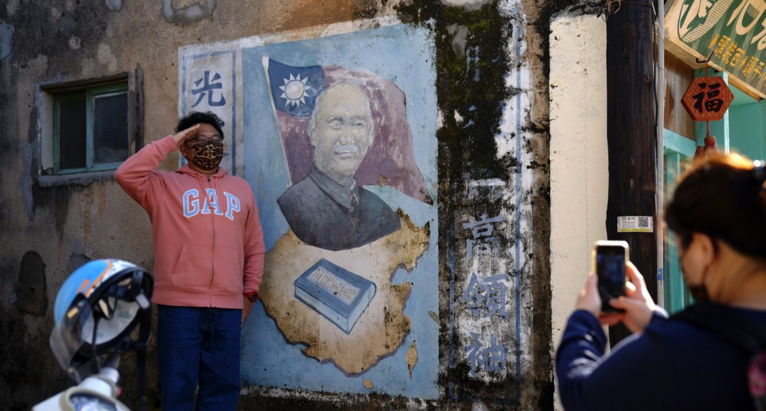Person with short curly hair and a GAP jacket salutes for a photo next to a faded street mural of a man agains the Taiwanese flag