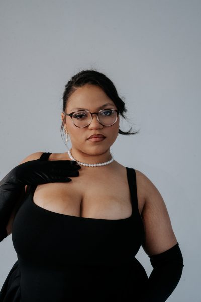 A woman wearing glasses, a black dress, black gloves, and a pearl necklace poses confidently against a plain light background, channeling the artistic spirit of Harborwalk Sounds ICA Berklee.
