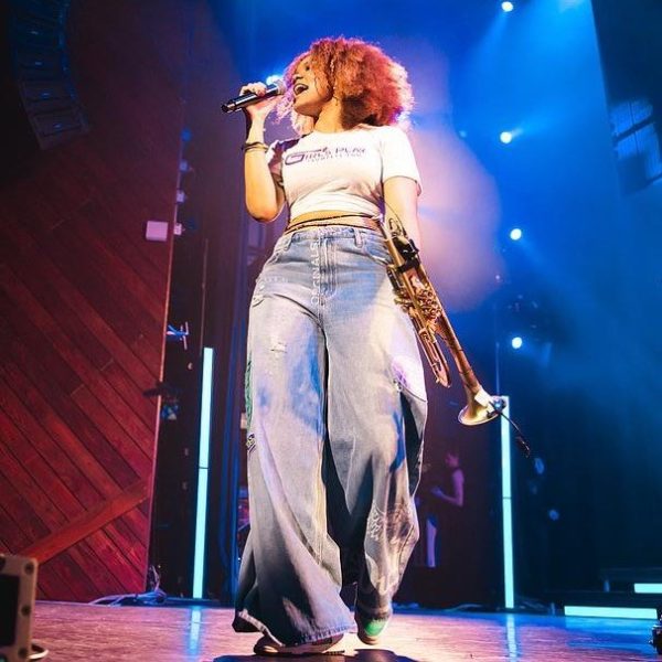 A woman with curly hair performs on stage at Harborwalk Sounds, holding a microphone and a trumpet. She wears a white crop top and wide-legged jeans, illuminated by colorful lights, showcasing Berklee talent.