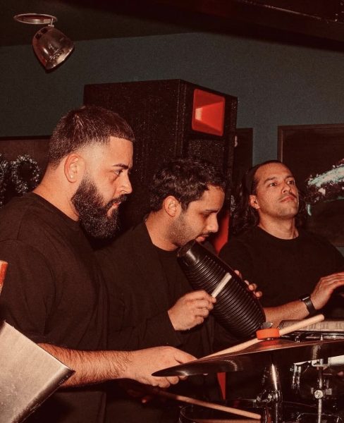 Three musicians dressed in black play percussion instruments, including a guiro and bongos, in a dimly lit indoor setting with a red-tinted light—capturing the vibrant energy of Vinyl Nights DJ events at ICA Boston.