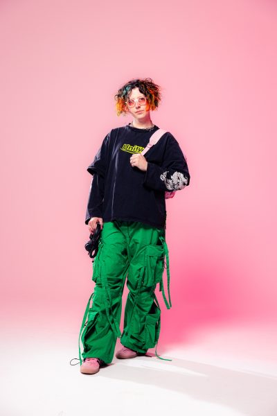 A person with curly, colorful hair wears pink sunglasses, a black long-sleeve shirt, bright green cargo pants, and pink shoes, standing against a pink background and holding a pink bag—ready for vinyl nights at ICA Boston.