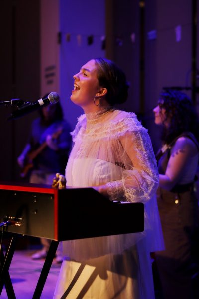 A woman in a white lace dress sings passionately into a microphone while playing a keyboard on stage at Harborwalk Sounds ICA, with other Berklee musicians performing in the background.