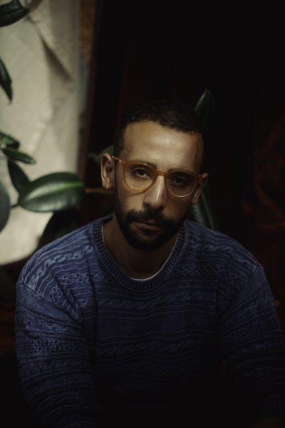 A man with short hair and a beard, wearing large orange glasses and a blue patterned sweater, sits indoors near a leafy plant with soft light and dark background—capturing the cool vibe of vinyl nights at ICA Boston.