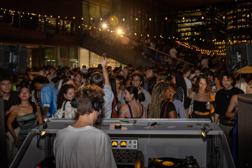 A DJ spins at an outdoor night party, energizing the crowd under glowing string lights. City buildings frame the scene as people enjoy the lively atmosphere