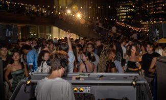 A DJ spins at an outdoor night party, energizing the crowd under glowing string lights. City buildings frame the scene as people enjoy the lively atmosphere