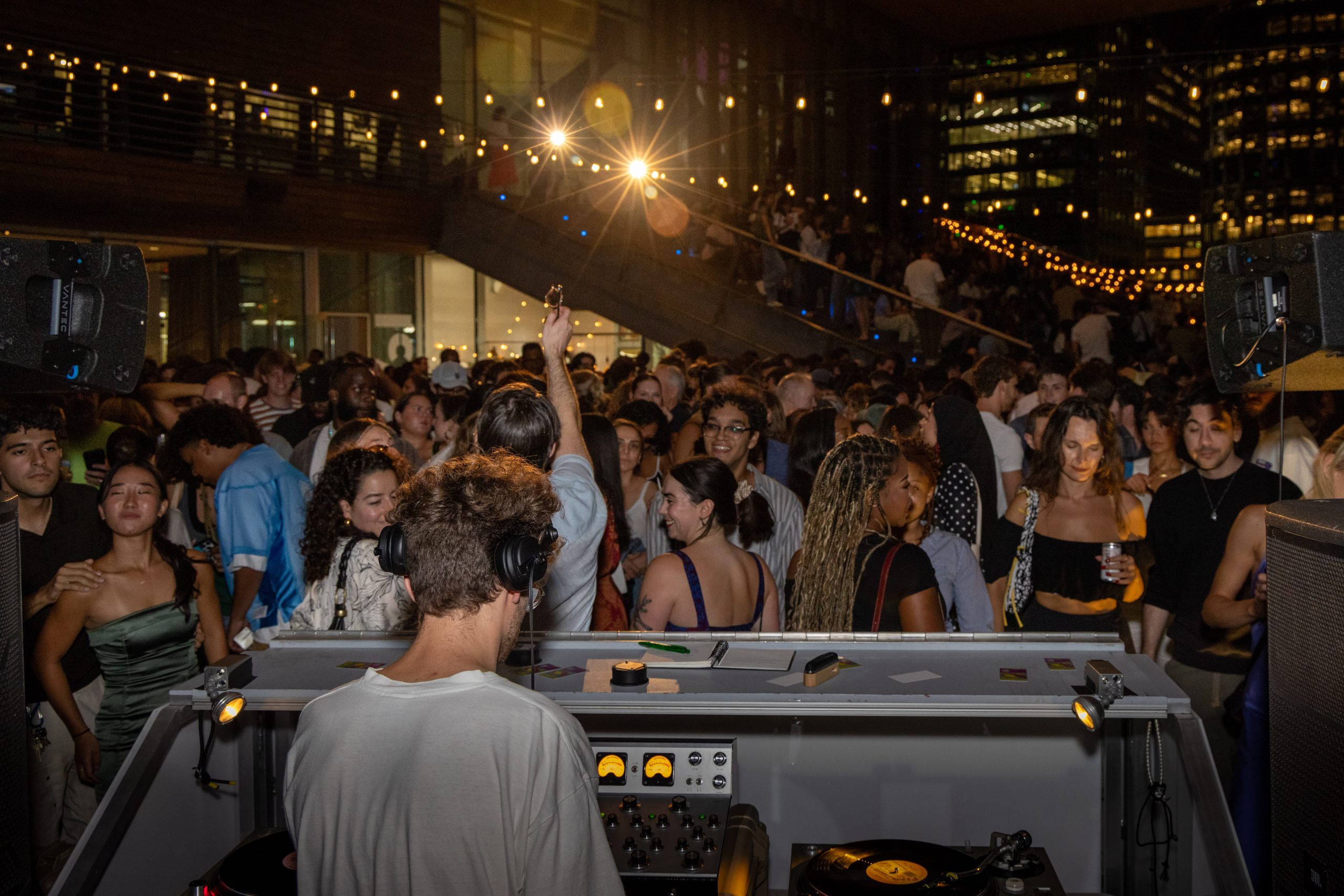 A DJ spins at an outdoor night party, energizing the crowd under glowing string lights. City buildings frame the scene as people enjoy the lively atmosphere