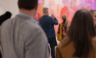 a man and a womans shoulders in the foreground frame another man and a woman who look at a large rectangular painting of red figures the man takes a photo of the work