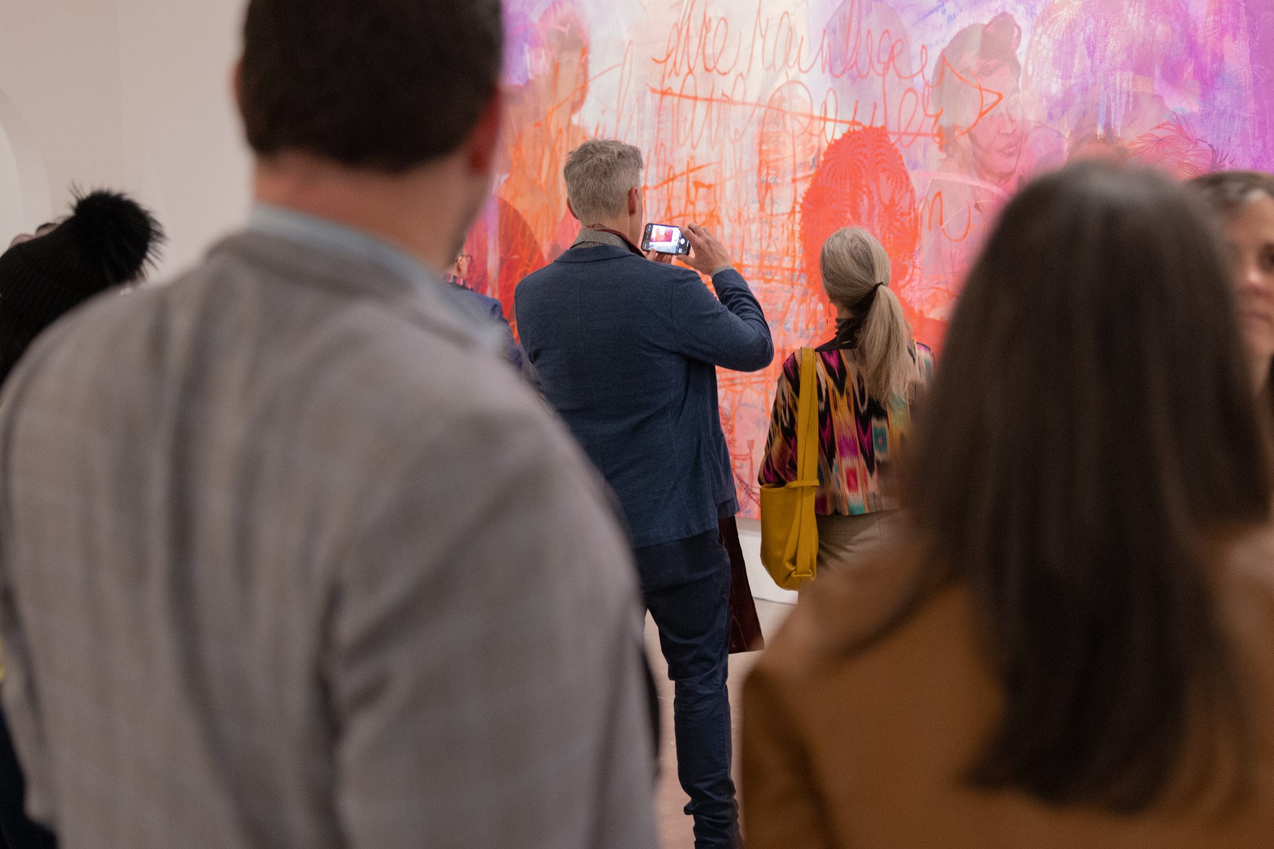 a man and a womans shoulders in the foreground frame another man and a woman who look at a large rectangular painting of red figures the man takes a photo of the work