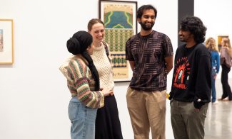 Four people stand together in an art gallery, smiling and talking. Framed artwork hangs on the white wall behind them.