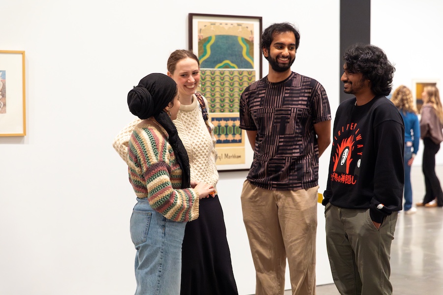 Four people stand together in an art gallery, smiling and talking. Framed artwork hangs on the white wall behind them.