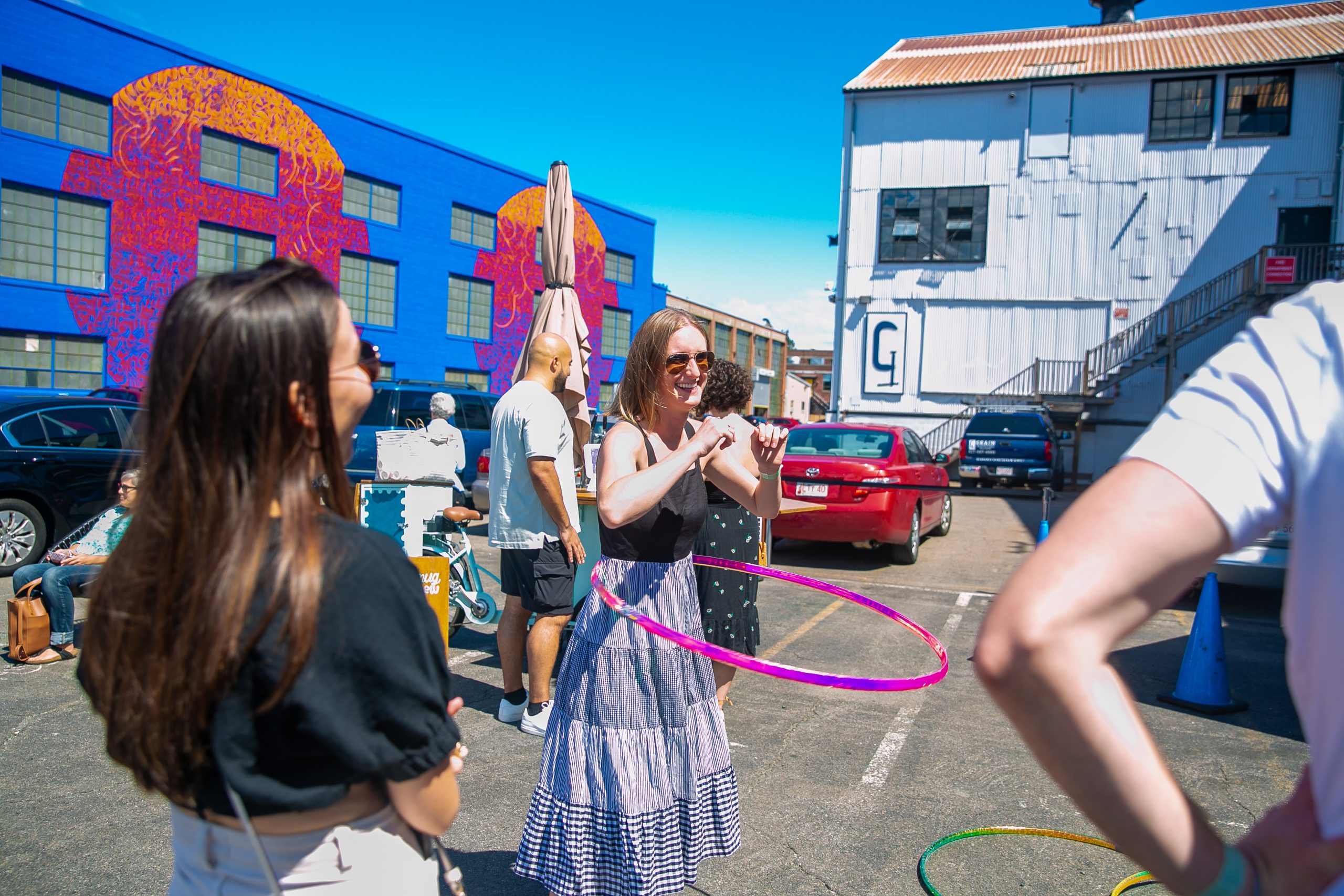 A woman in sunglasses hula hoops in a sunny parking lot, surrounded by people at the ICA Boston Watershed block party. Colorful street art and a white building are in the background.