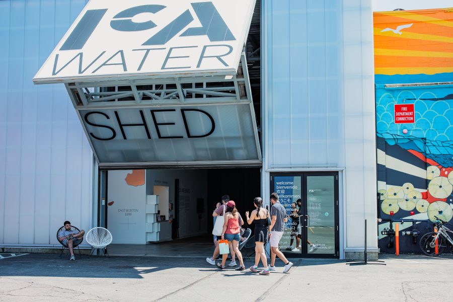 A group of people walking into the ICA Watershed building, featuring a large open entrance, clear signage, and colorful murals on the exterior wall. A person sits on a chair outside.