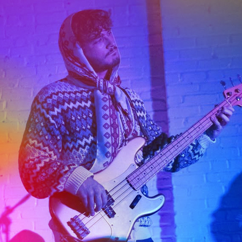 A person wearing a patterned sweater and scarf plays an electric bass guitar in front of a brick wall, with colorful lighting creating a vibrant, Harborwalk Sounds ICA Berklee atmosphere.
