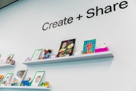 A wall labeled "Create + Share" with shelves of artworks of varying media on display, including clay vessels, drawn portraits, and a two dimensional work with fried eggs