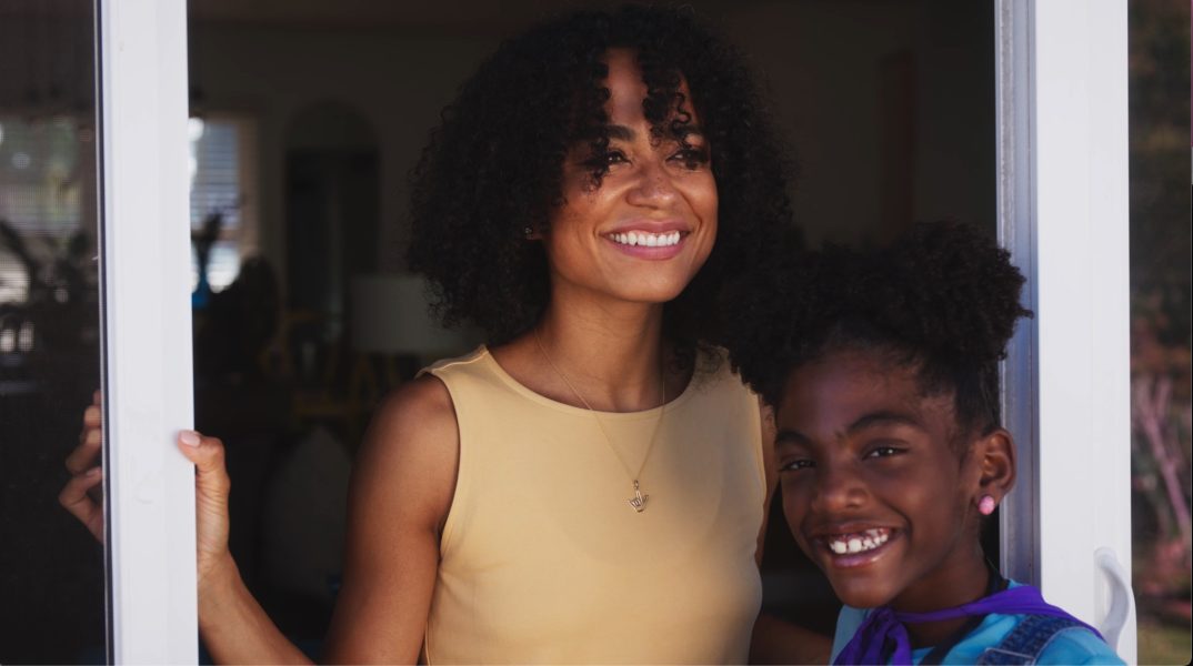 A smiling Black parent with shoulder length hair and their child by a doorway