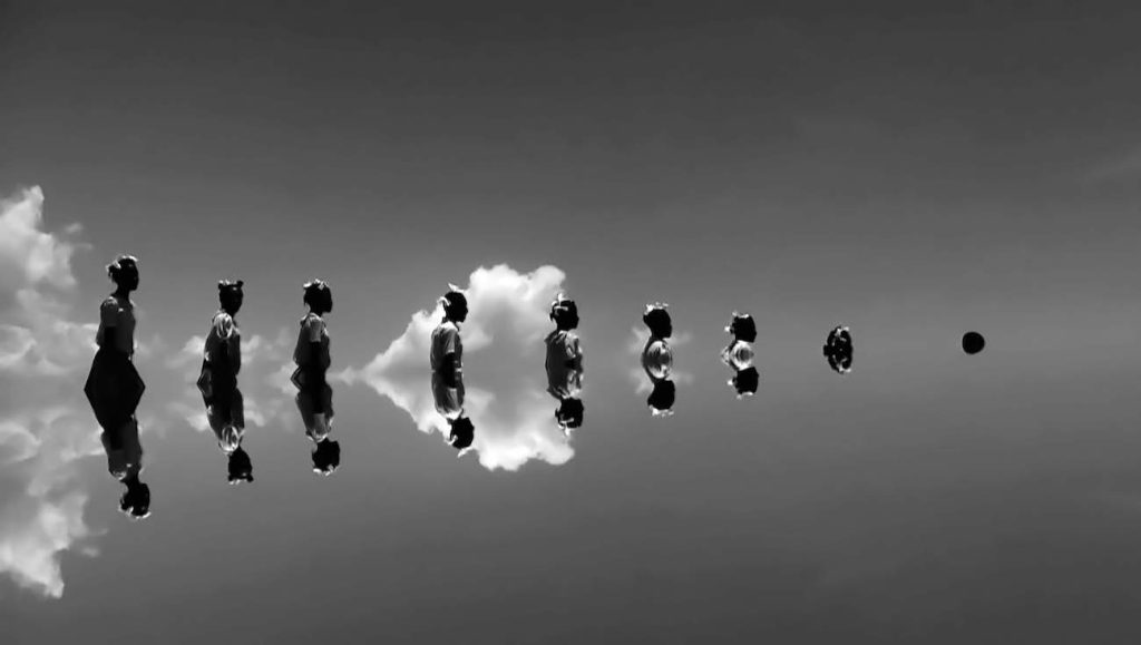 A black and white photo of a line of 9 figures reflected in water, increasingly submerged.