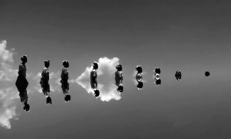 A black and white photo of a line of 9 figures reflected in water, increasingly submerged.