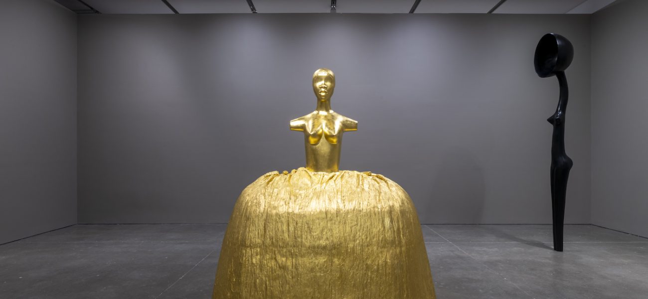 Two sculptures in a room with gray walls: a gold female figure with a voluminous skirt and an elongated, stylized female bronze with a satellite dish in place of a head. 