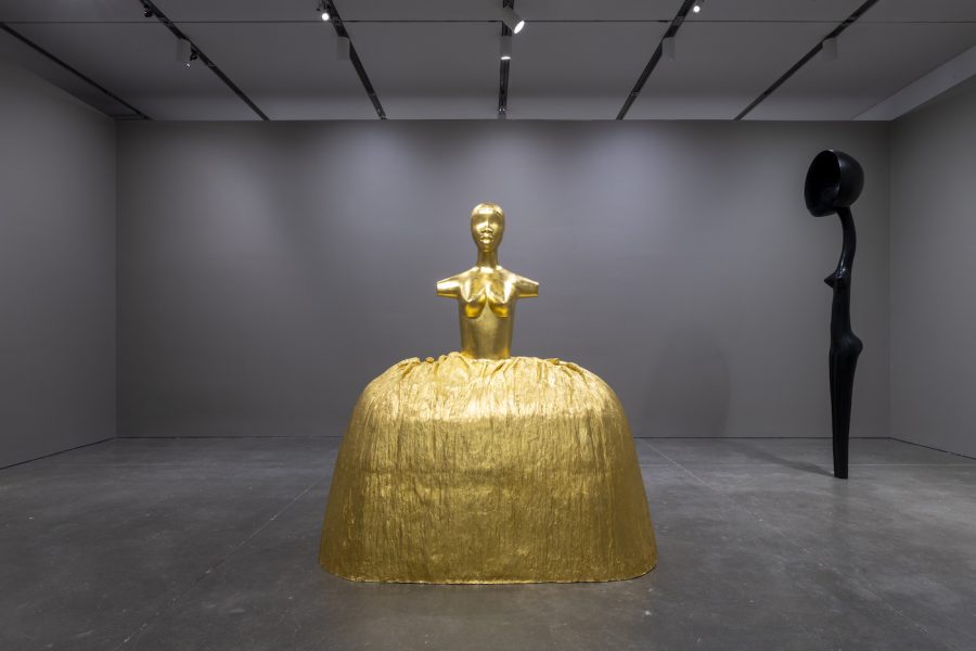 Two sculptures in a room with gray walls: a gold female figure with a voluminous skirt and an elongated, stylized female bronze with a satellite dish in place of a head. 