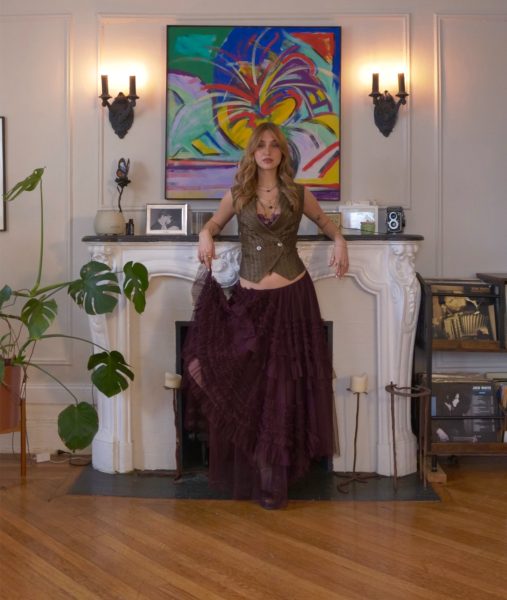 A woman in a long, tiered purple skirt and brown vest stands in front of a white fireplace, with colorful abstract art and vintage decor on the mantel, as Harborwalk Sounds from Berklee play softly in the stylish living room with wooden floors and houseplants.