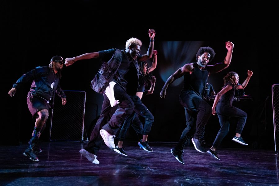 a group of dancers in all black and sneakers are mid air in a lunge