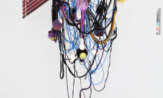 Photograph of sculpture made from a clothing rack draped in cable, rope, cord, light bulbs and aluminum blinds.