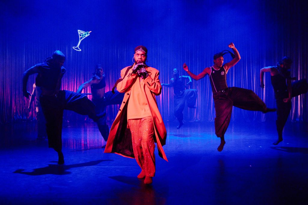 A person singing into a microphone and dancers in the background.