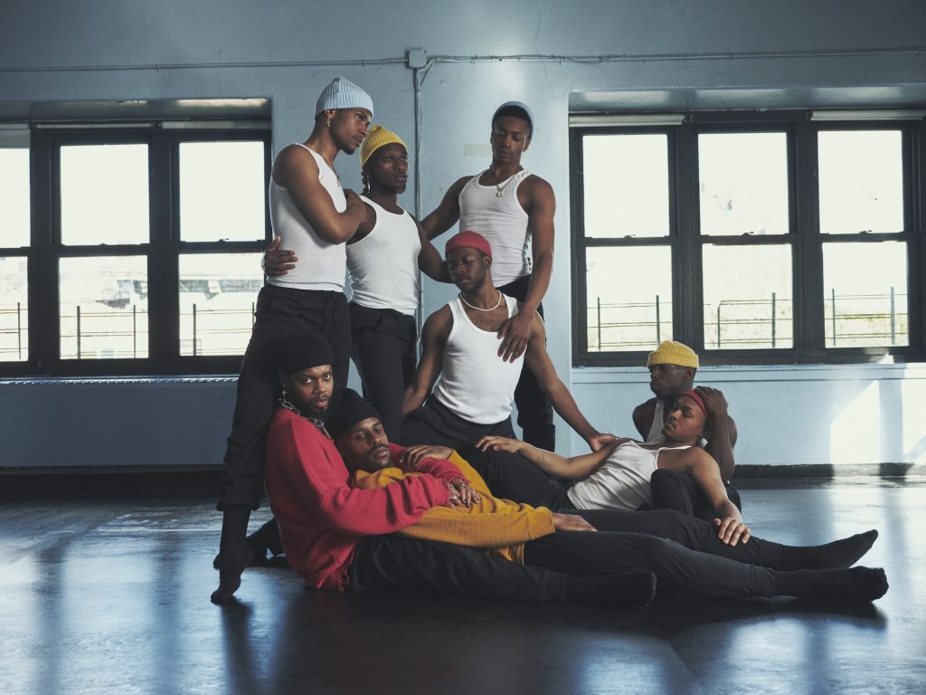 Men wearing white tank tops and beanies intertwined together on a dance floor