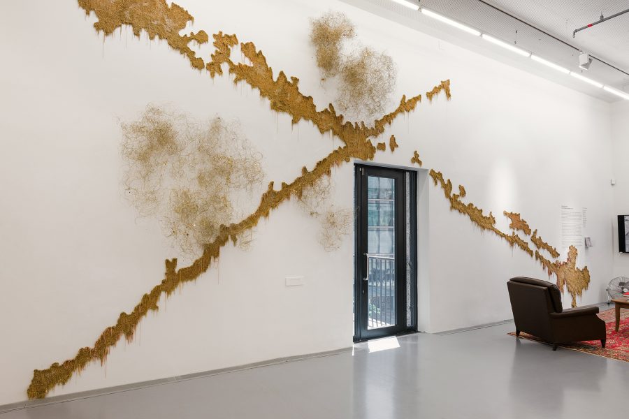 A large room with white walls and an artwork consisting of a large, camel-colored, X-shaped form reaching over a doorway, plus suspended cloudlike forms.