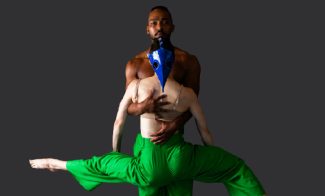 Performer in blue headdress and green pants is embraced by a performer behind them