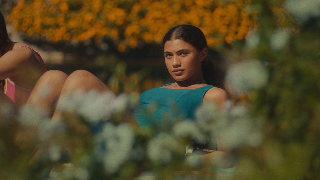 A young woman wearing a blue swimsuit reclines outdoors, surrounded by blurred greenery and vibrant yellow flowers in the background, gazing directly at the camera—her pose reminiscent of portraits showcased at ICA Boston.