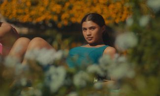 A young woman wearing a blue swimsuit reclines outdoors, surrounded by blurred greenery and vibrant yellow flowers in the background, gazing directly at the camera—her pose reminiscent of portraits showcased at ICA Boston.