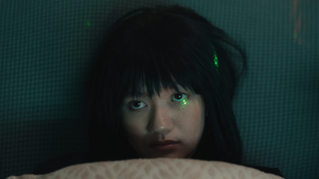 An Asian woman looks up from under a pink blanket with green light speckled on her face. Still from When You Left Me On That Boulevard.