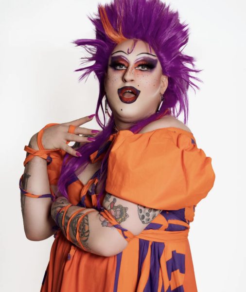 Coleslaw has purple hair and is wearing an orang dress in a white room