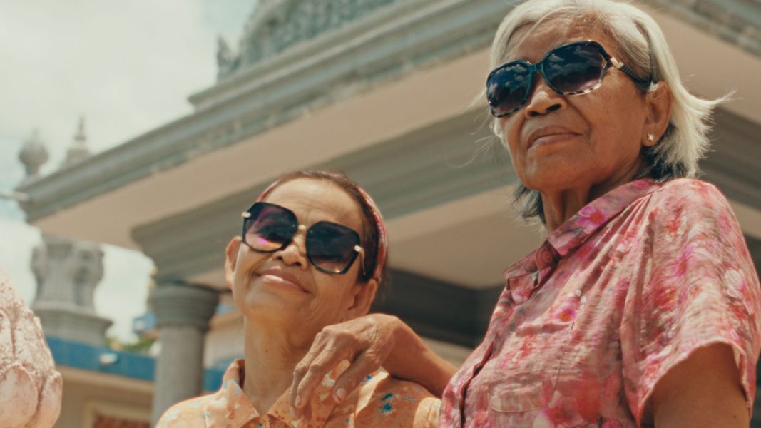 Two older women wearing sunglasses and colorful shirts stand outside, smiling and posing confidently in front of a building with columns and detailed architecture, reminiscent of portraits taken at the Sundance Film Festival or ICA Boston.