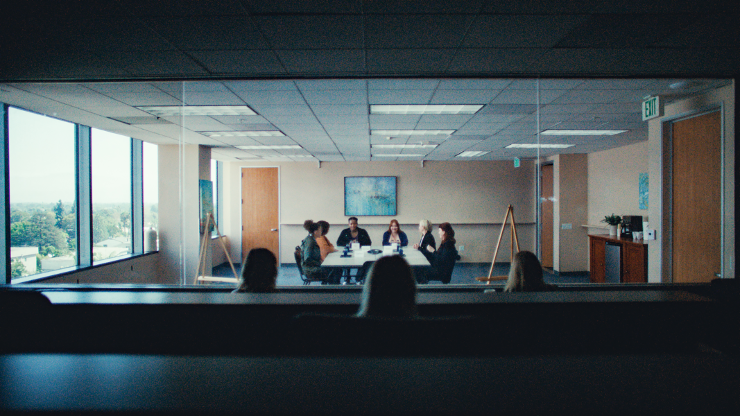 Two women in a dimly lit office space watch a focus group through a glass wall. The focus group is made of 6 women who all sit at a rectangular table in a brightly lit room with beige colored walls and tan doors.