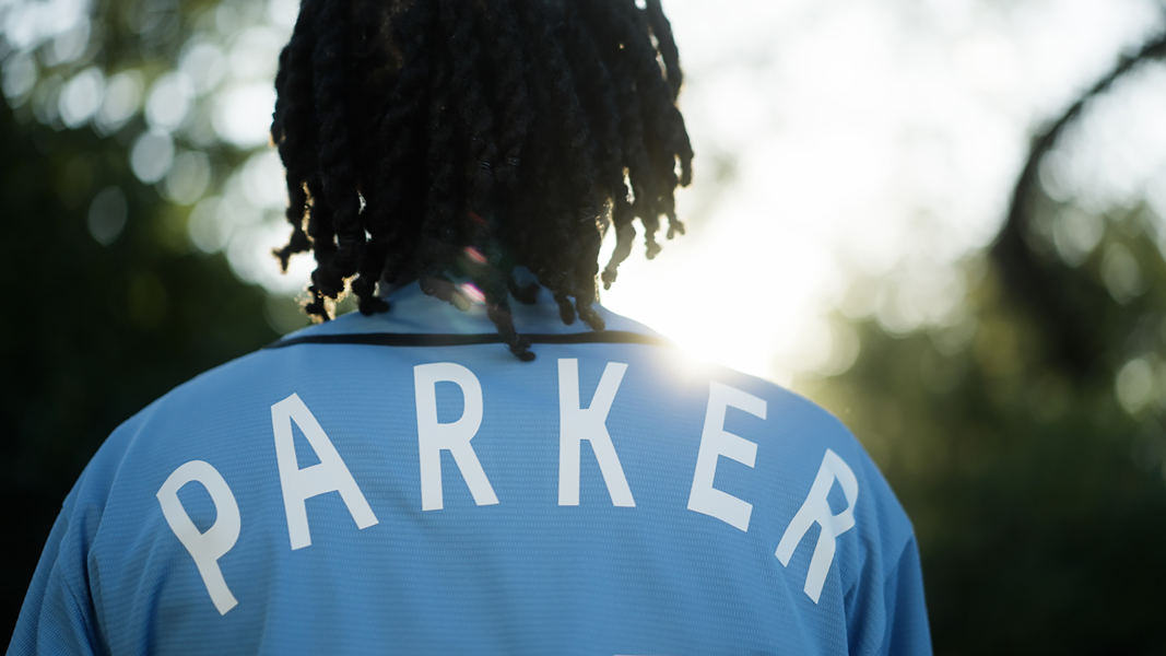 The of a man with black braided hair wears a blue jacket that says parker and looks onto greenery that is out of focus.