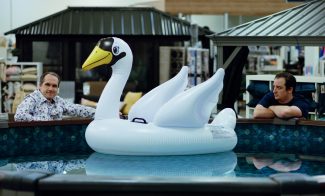 Two men visible only form the shoulders up stand at about at a short distance from one another outside of an above ground pool with a large white blow up swan in it.