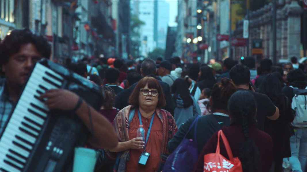 A middle-aged woman with glasses stands in a crowded city street, holding a water bottle—her calm presence reminiscent of scenes from the Sundance Film Festival or ICA Boston. Nearby, a man plays an accordion as people walk in different directions around her.