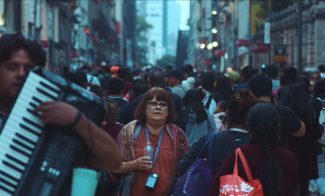 A middle-aged woman with glasses stands in a crowded city street, holding a water bottle—her calm presence reminiscent of scenes from the Sundance Film Festival or ICA Boston. Nearby, a man plays an accordion as people walk in different directions around her.
