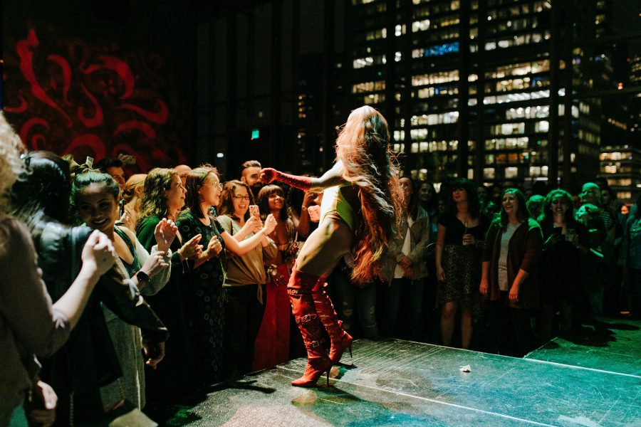 Person with long blonde hair wearing a white body suit and red boots dances at the edge of the stage while a crowd watches and dances along.