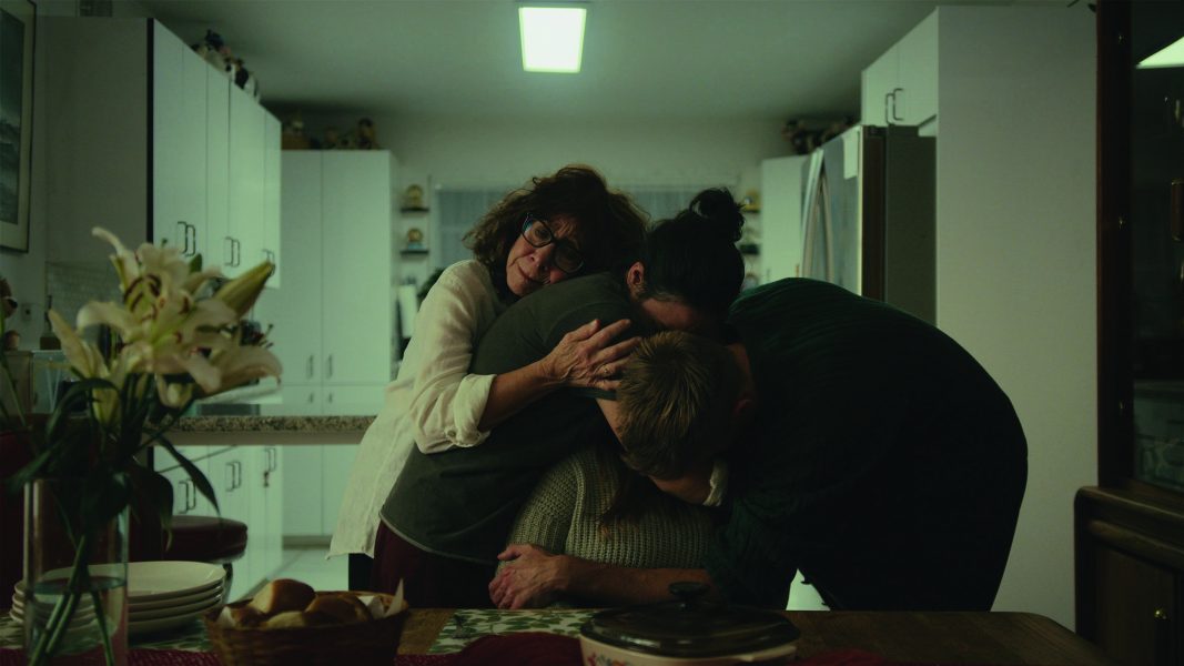 Three people embrace closely in a brightly lit kitchen, comforting each other. With white cabinets and flowers on the table, the emotional scene feels reminiscent of an indie film screening at Sundance Film Festival or ICA Boston.