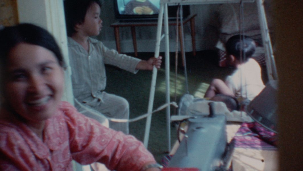 A woman smiles at the camera near a sewing machine, while two young children—one on the floor and one watching a cartoon—fill the cozy, homey scene. It’s a moment worthy of Sundance Film Festival or ICA Boston storytelling.
