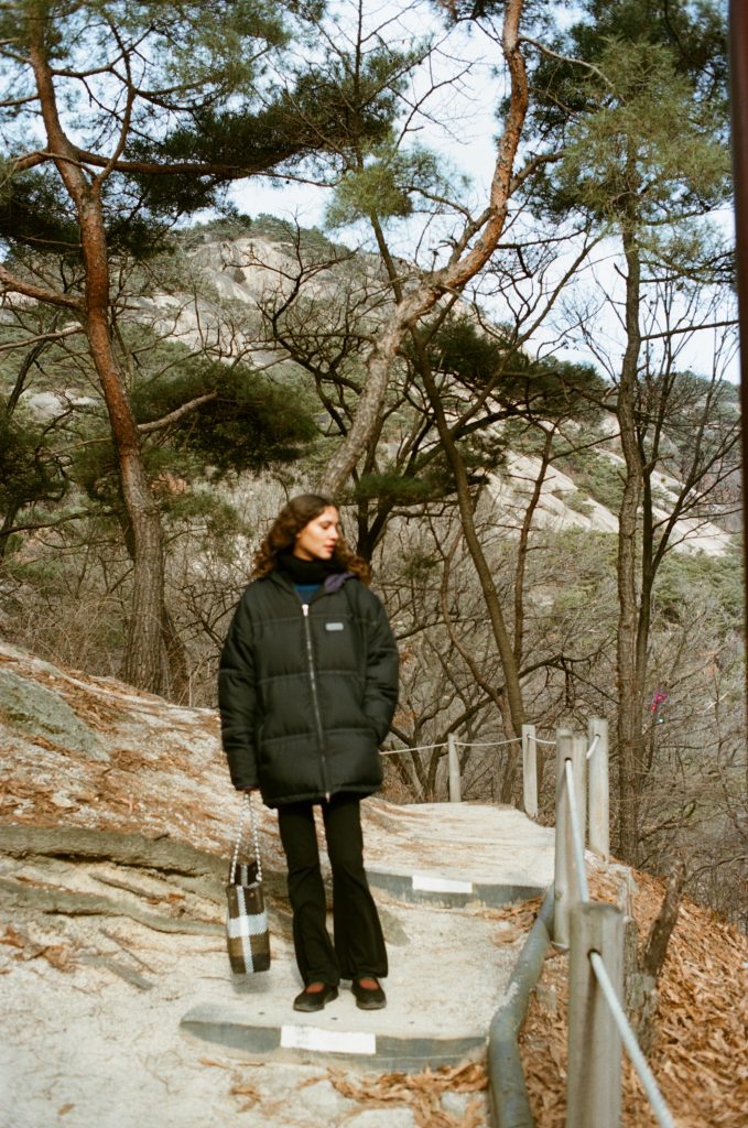 An individual with long dark hair in a black puffer jacket and black leggings stands on a rock in a forest