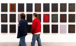 Two people stand in front of a wall displaying a grid of abstract, mostly dark paintings with geometric designs. Both wear casual clothes and are looking at the art pieces.