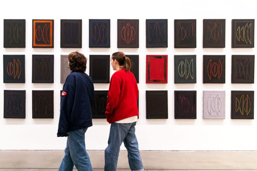 Two people stand in front of a wall displaying a grid of abstract, mostly dark paintings with geometric designs. Both wear casual clothes and are looking at the art pieces.