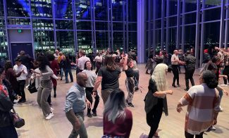 around 50 people simile and dance in a room lit by overhead lights, because it is night time. the room has floor to ceiling windows that overlook other buildings visible only by the light from their windows
